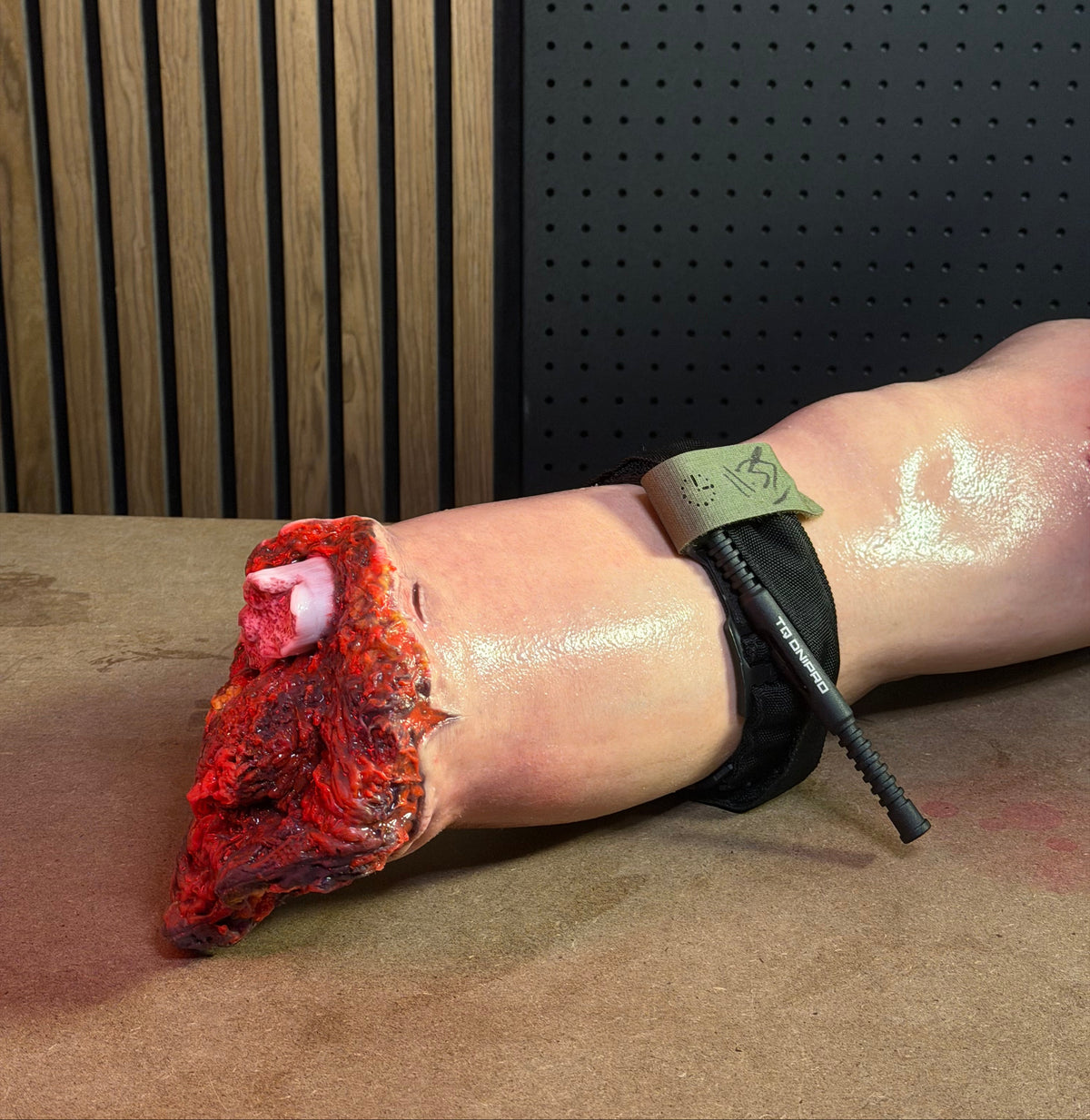 A close-up of a simulated lower leg and ankle with a black tourniquet applied, with a realistic bloody amputation. The background shows a dark perforated panel and wood slats.