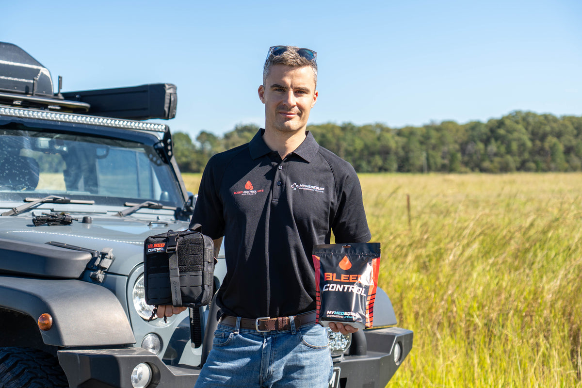 MyMedEquip's (GREMED GROUP) Director Theo, standing in front of a 4wd in the countryside. He is holding a Tactical Trauma Kit in one hand and a Public Access Bleed Control Kit in the other. The kits and his shirt and Black with Red Trim. It is a sunny blue skied day.