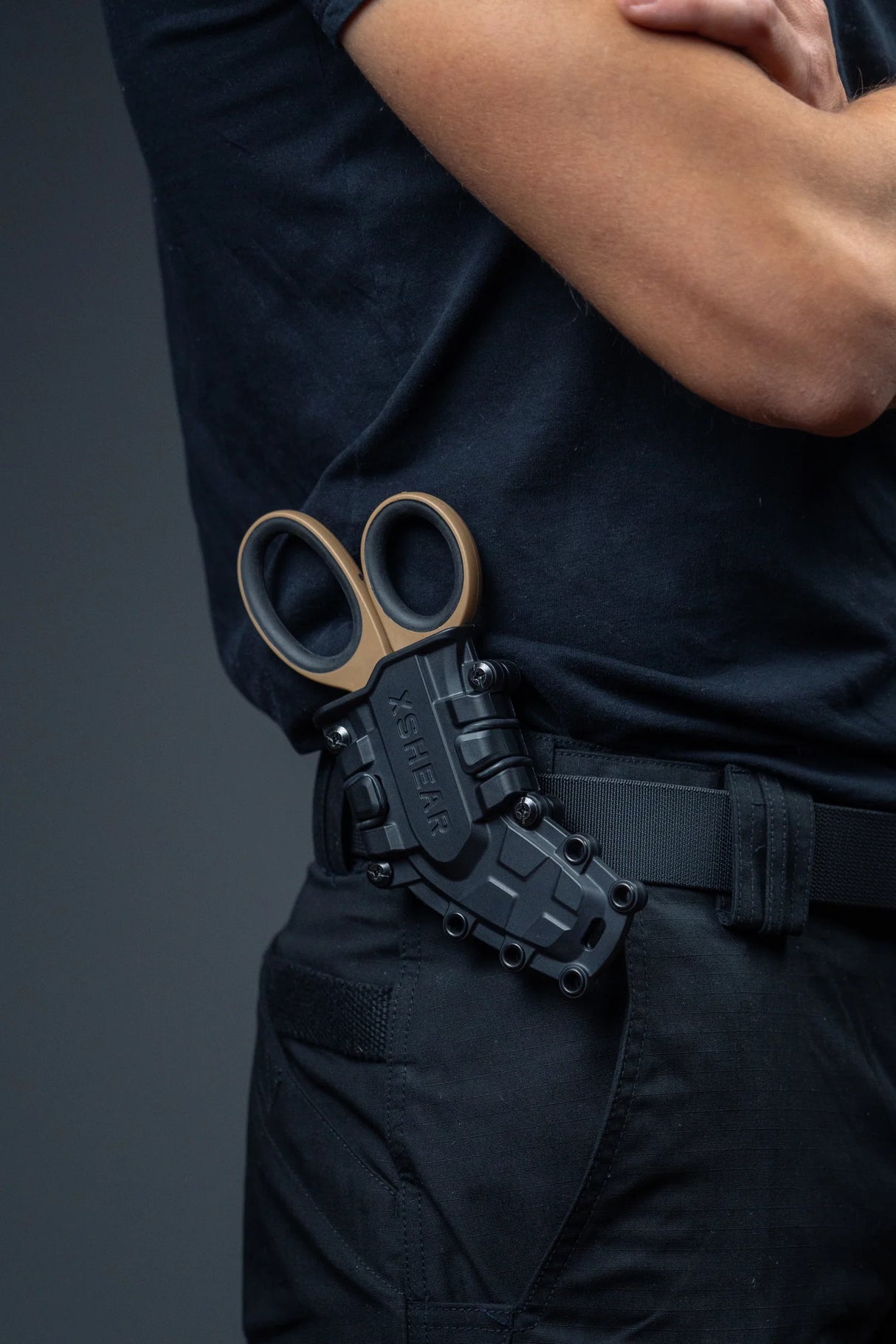Black tactical Trauma Shear holster made of molded ABS with multiple attachment points and retention bands, holding a set of coyote brown XShears and mounted on belt of person wearing navy clothing with a grey background.